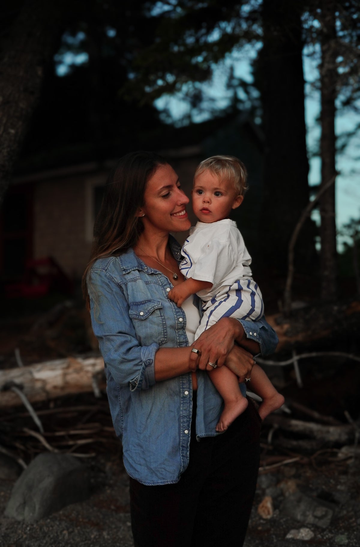 A mother holding her toddler outdoors, standing near trees.